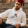 Arabic man wearing white color of Arabi hoodie for men with the Arabic word "عربي" in modern Arabic calligraphy design, and a classic red Tarboosh on his head