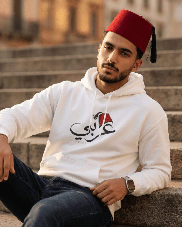Arabic man wearing white color of Arabi hoodie for men with the Arabic word "عربي" in modern Arabic calligraphy design, and a classic red Tarboosh on his head
