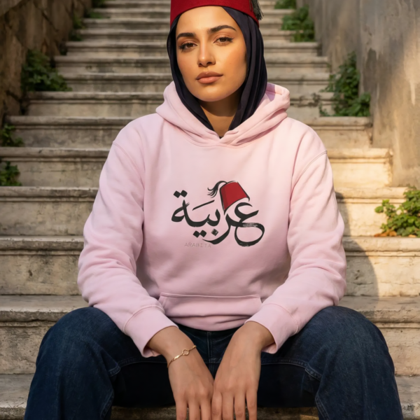 Arabic woman wearing black hijab and light pink color of Arabiya hoodie for women with the Arabic word "عربية" in modern Arabic calligraphy design