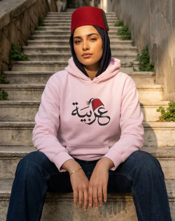 Arabic woman wearing black hijab and light pink color of Arabiya hoodie for women with the Arabic word "عربية" in modern Arabic calligraphy design